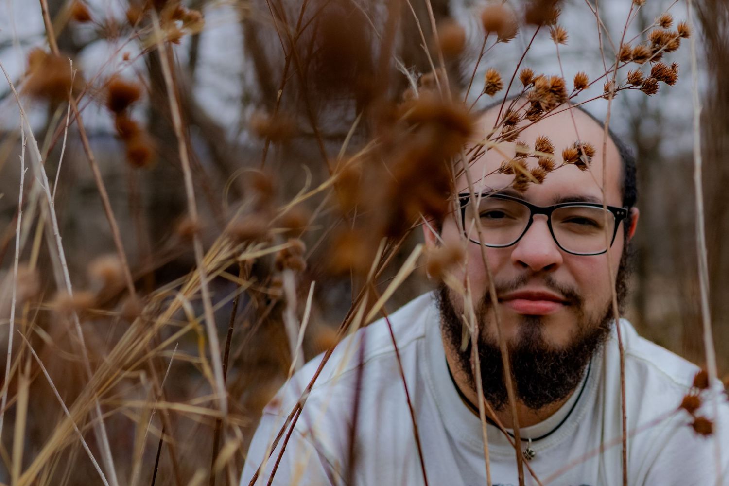 Close up autumn portrait of a person with glasses and a beard smiling among brown dried plants and grass.