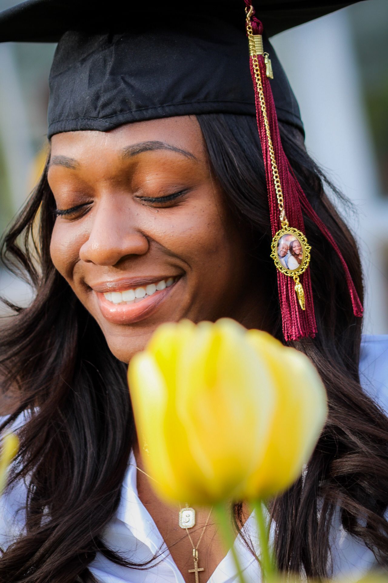 Graduate in cap and gown smiles peacefully with a yellow tulip in the foreground.