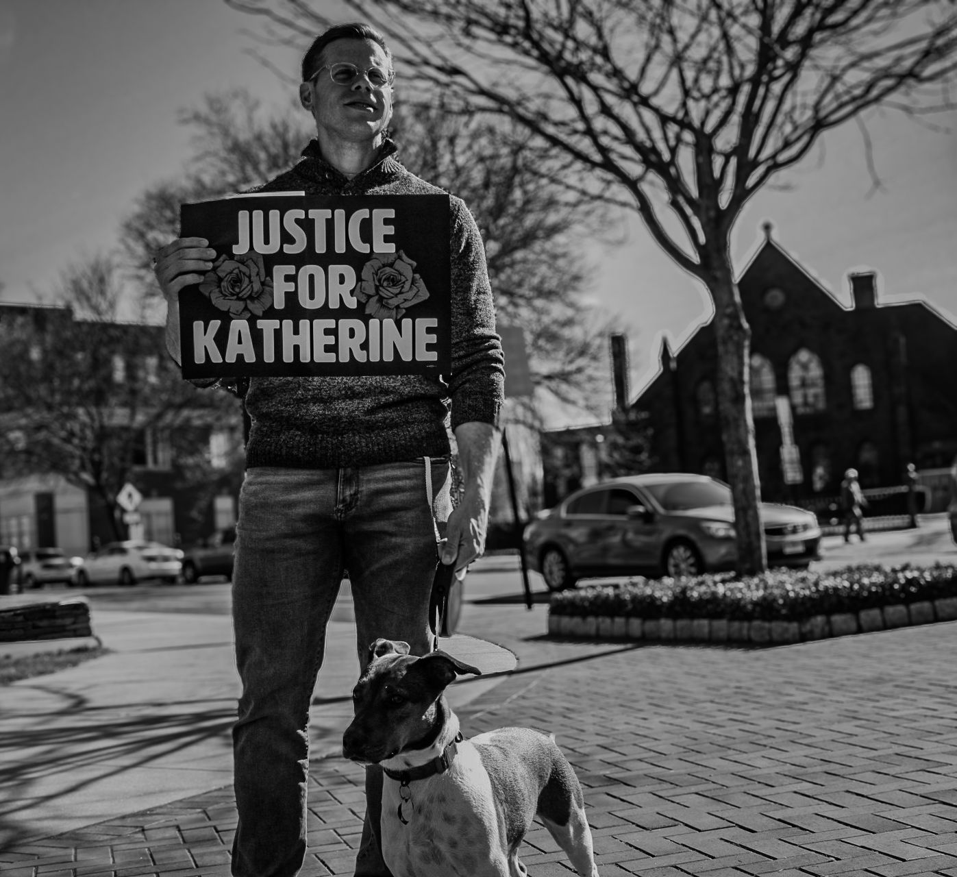 Person holding 'Justice for Katherine' sign stands with dog on brick sidewalk in black and white photo.