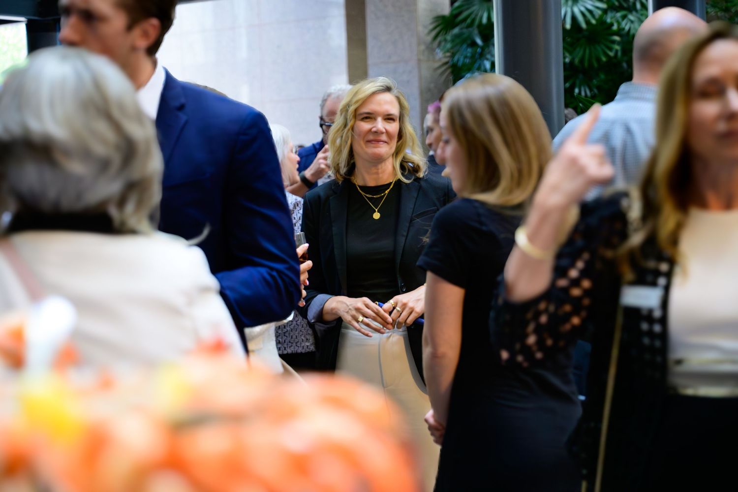 Woman in black dress smiling at an indoor networking event with guests mingling in a bright venue.