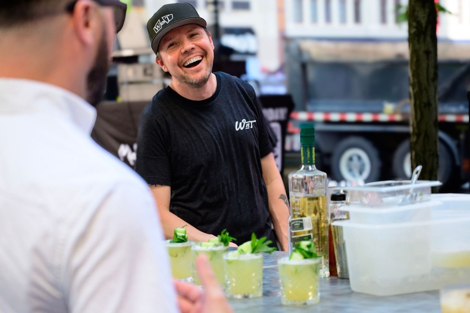 Smiling bartender in black shirt prepares cocktails with fresh herbs at an outdoor bar event.