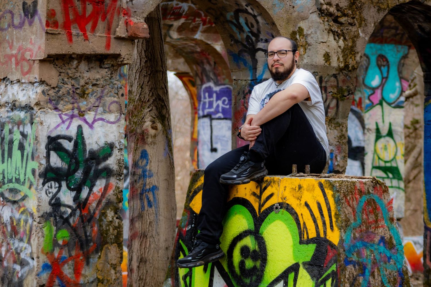 Person sits atop colorful graffiti wall in urban setting surrounded by vibrant street art and tags.