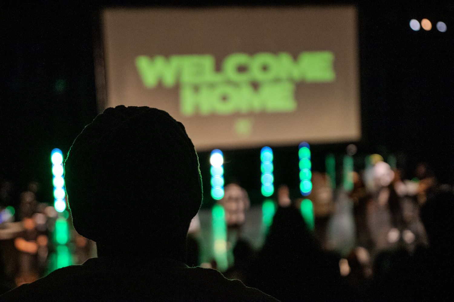 Silhouette of audience member watching stage with green Welcome Home sign and LED lights.