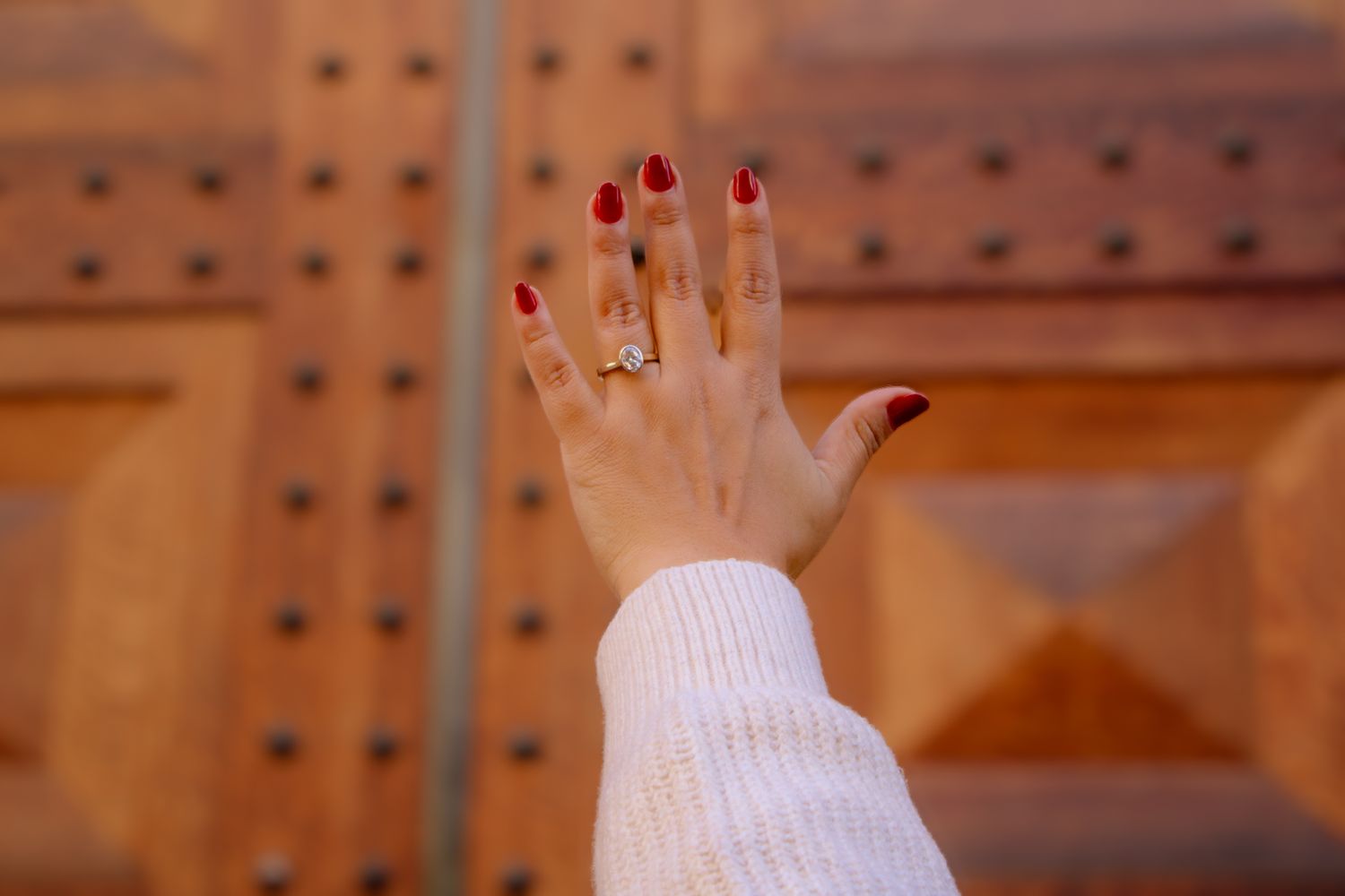 A hand wearing a white sweater displays a sparkling engagement ring against a wooden door.