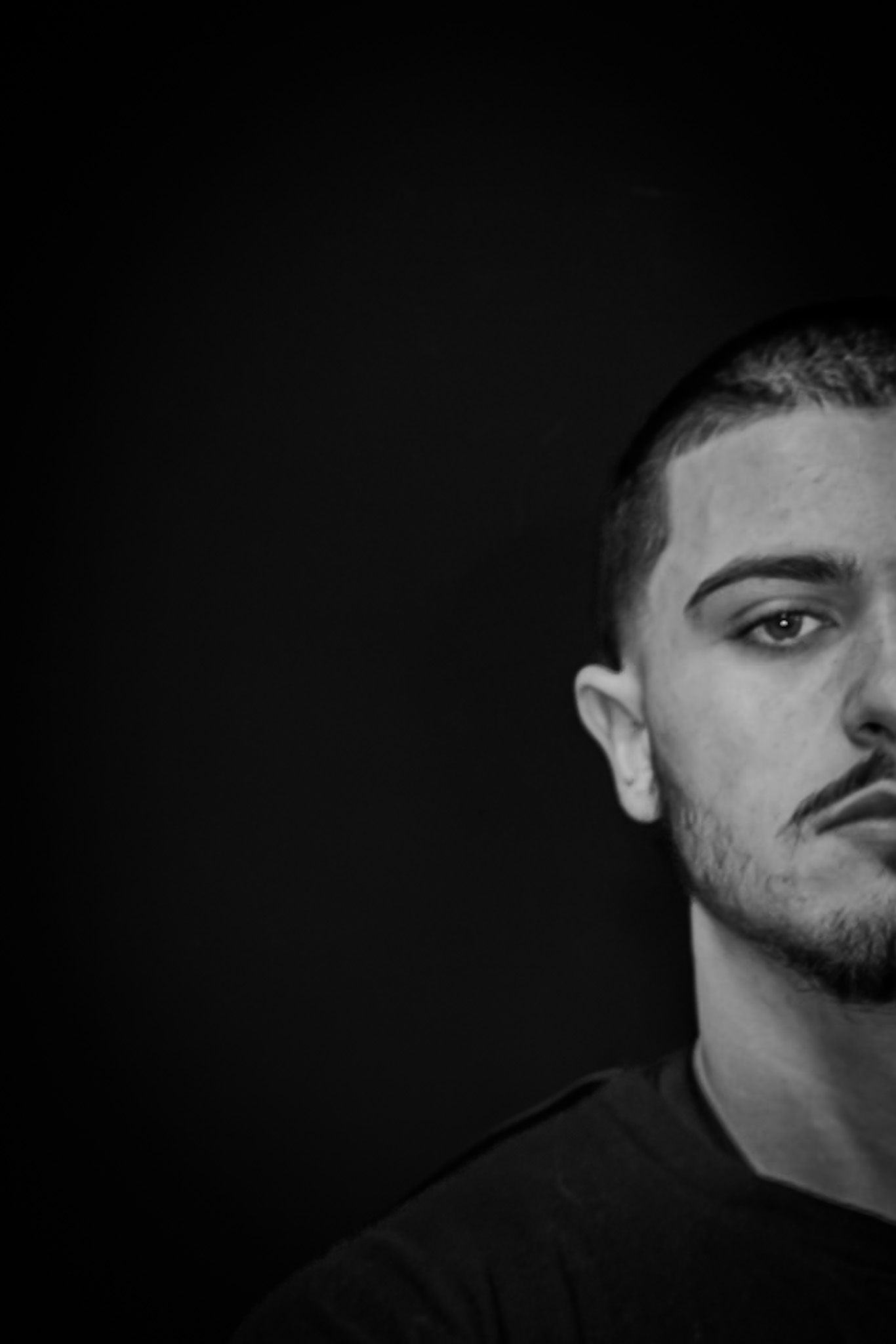 Dramatic black and white portrait against dark background shows half of subject's face.