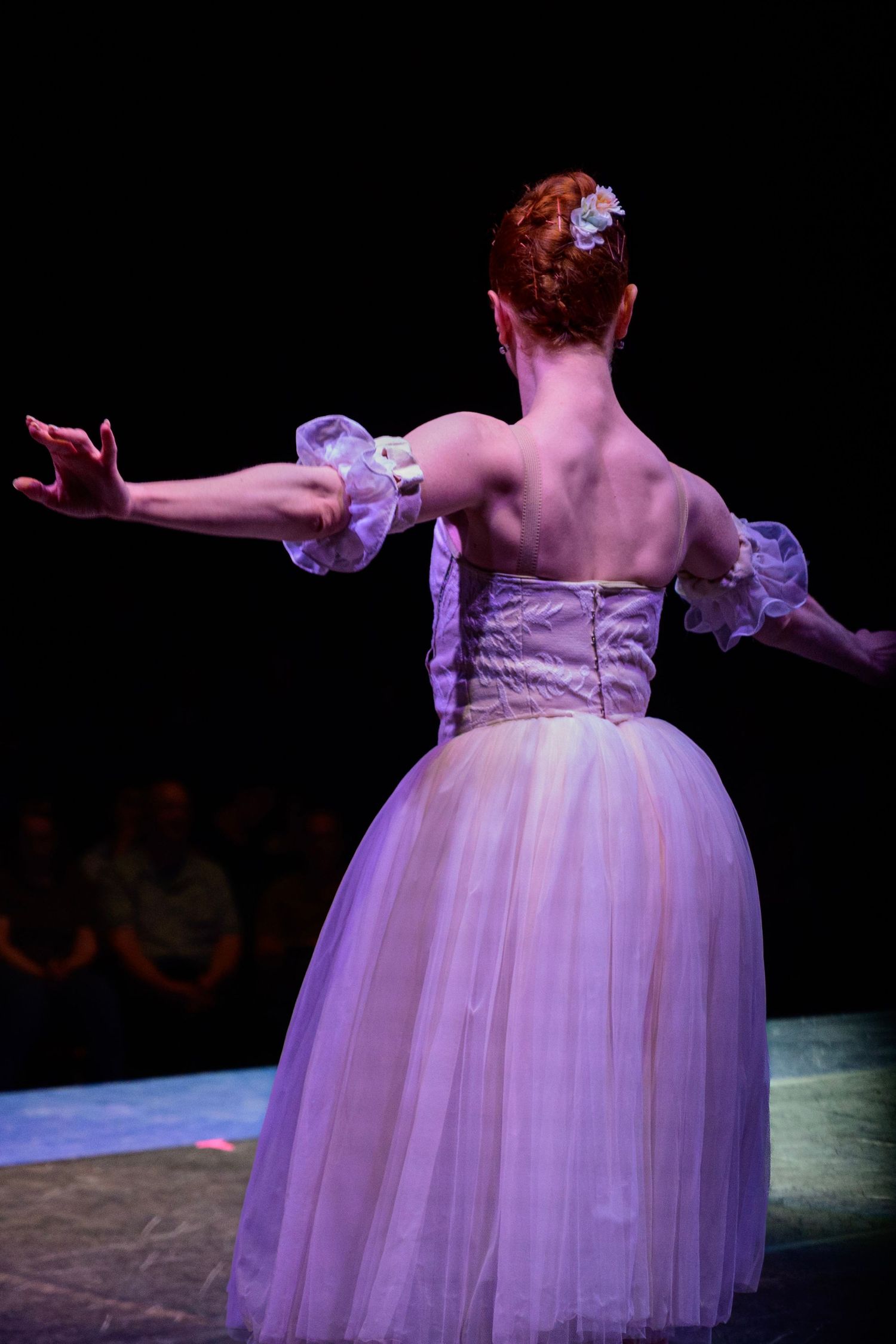 Red-haired ballerina in lavender tutu performs on stage with arms outstretched, viewed from behind.