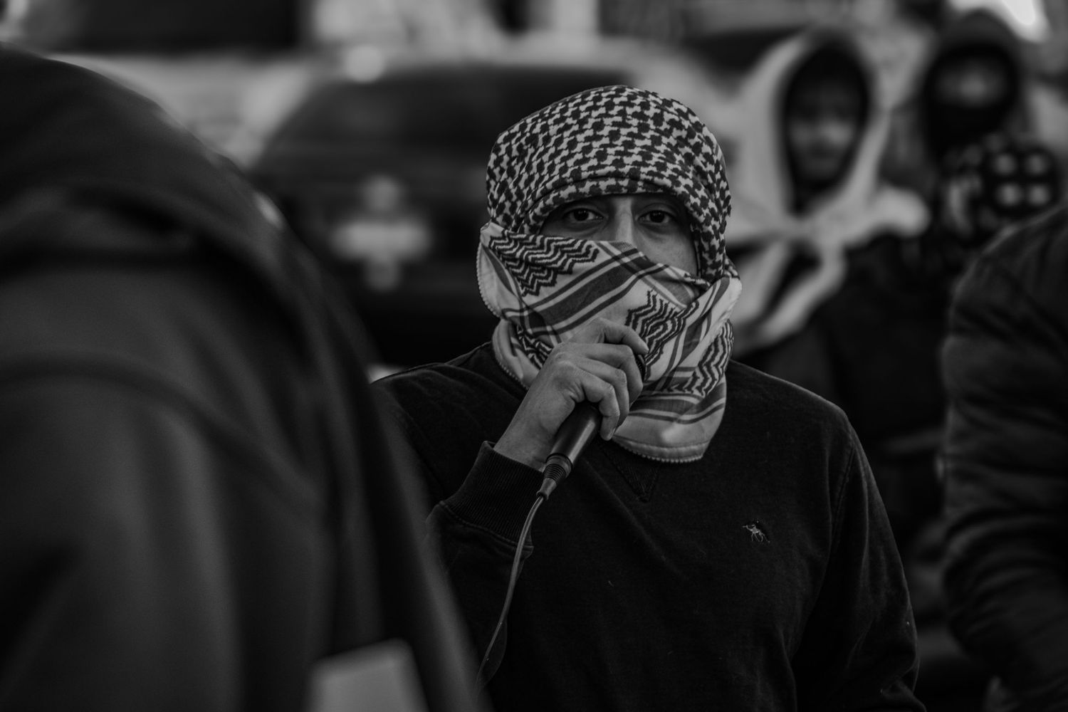 A person wearing a patterned keffiyeh head covering and sweater raises their hand to their face in a black and white photo.