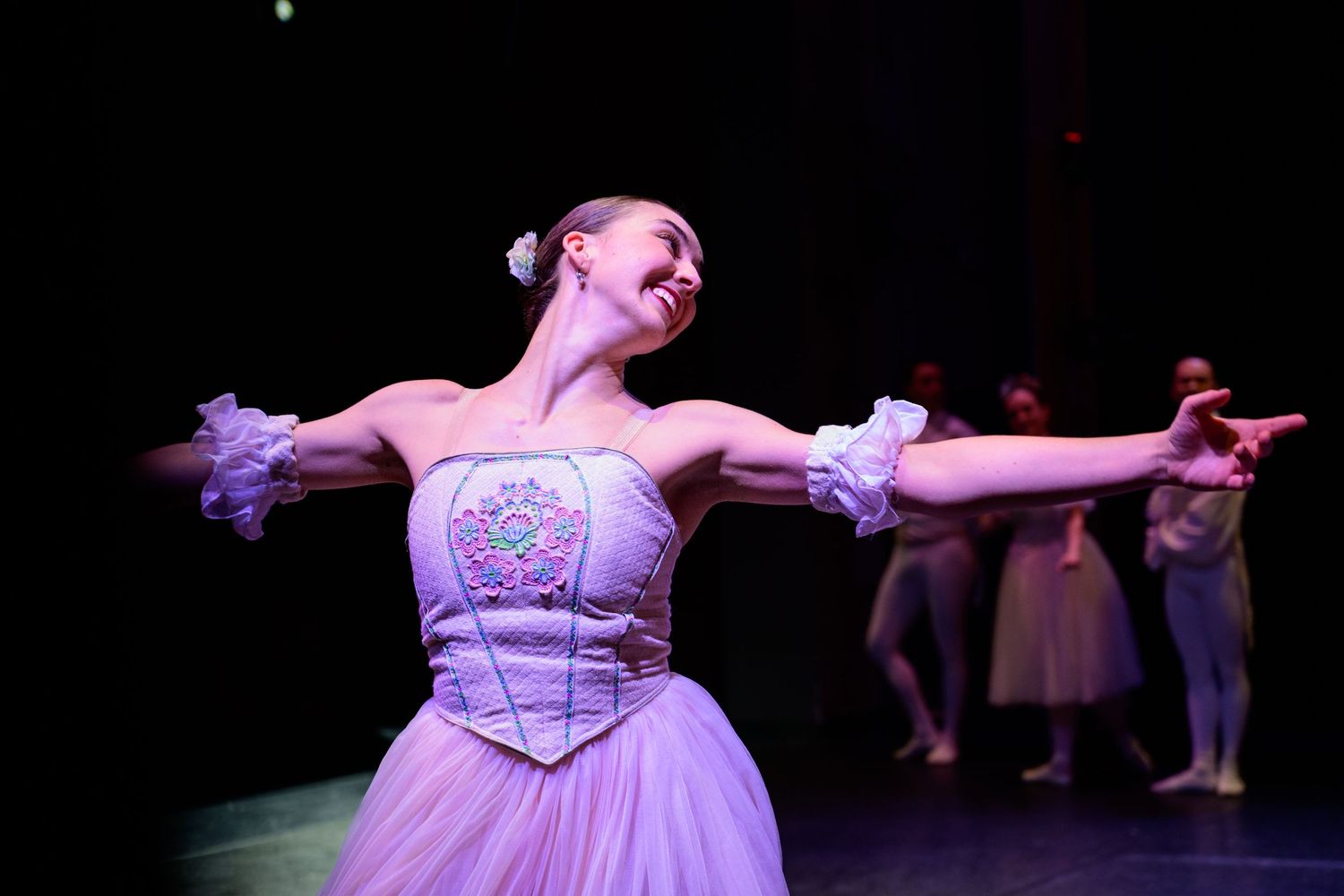 A smiling ballerina in a lavender tutu performs on stage with arms outstretched during a ballet performance.