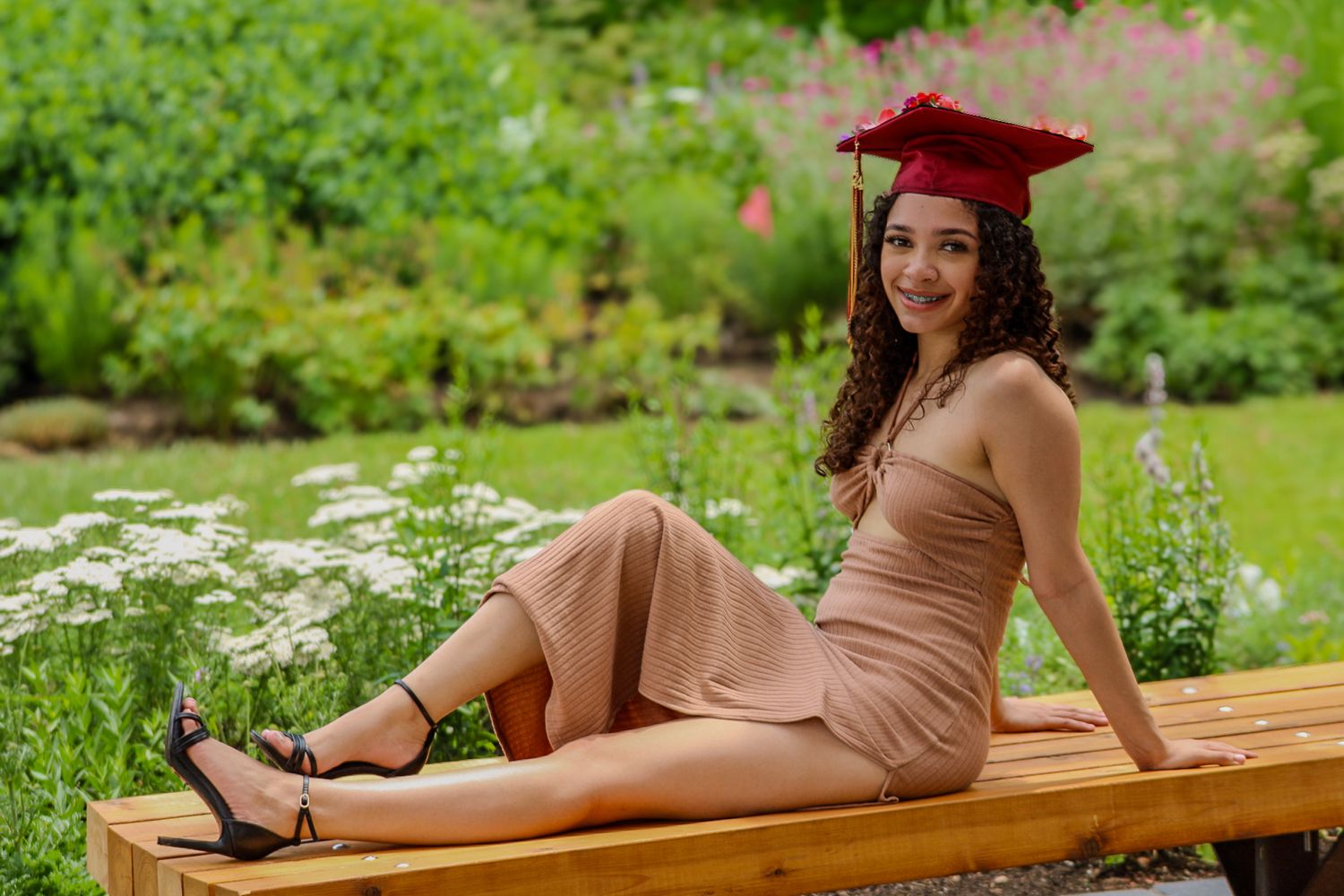 Graduate in red cap and beige dress poses on wooden bench in garden setting.