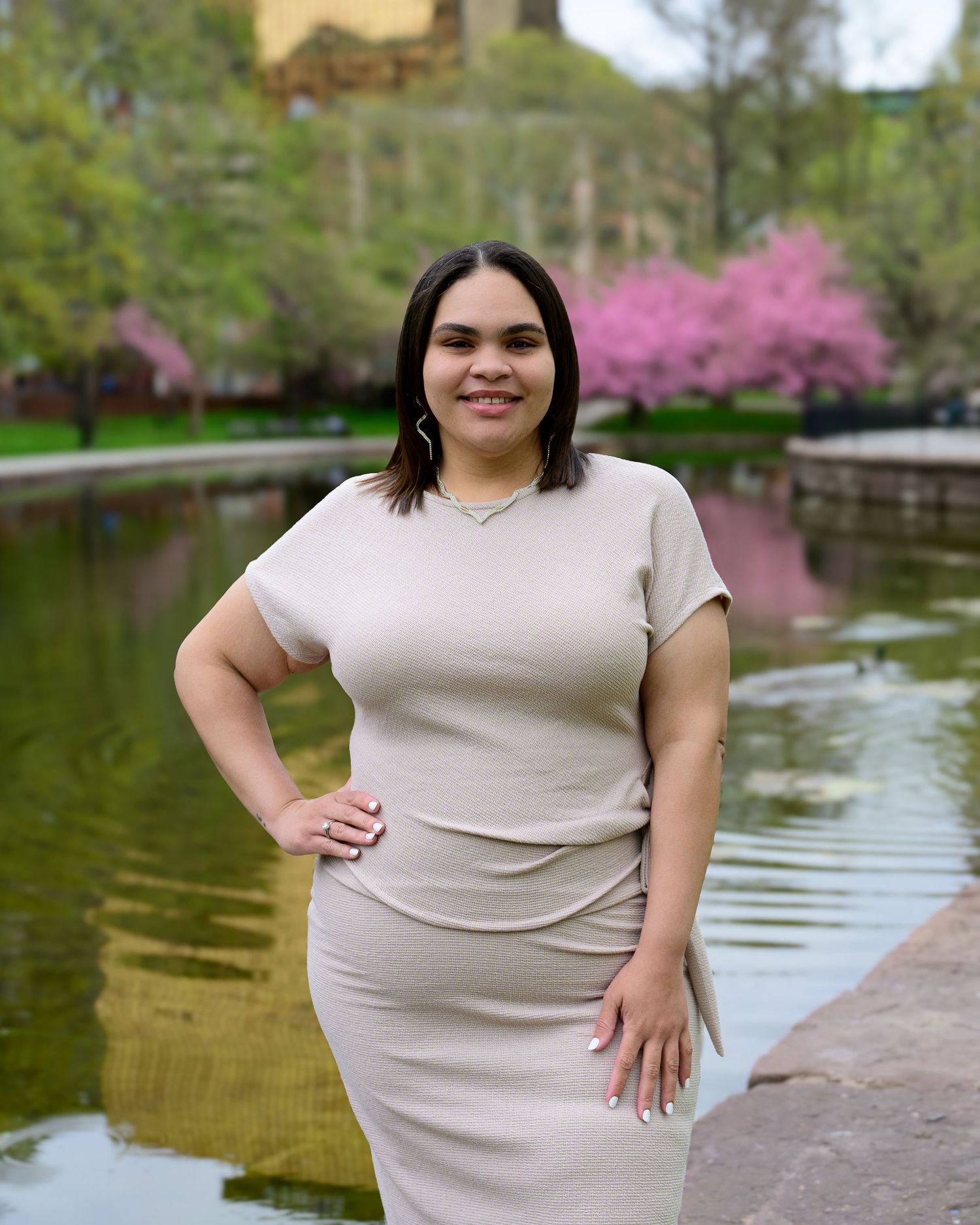A young woman in a beige dress poses by a pond with pink flowering trees in a park.