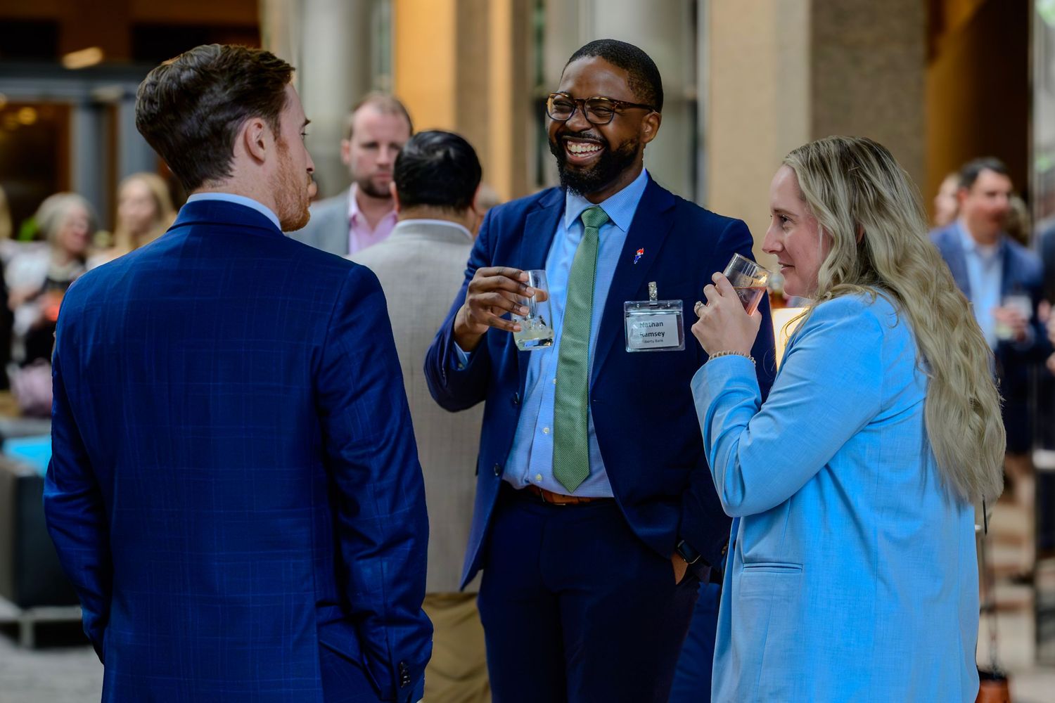 Three professionals in business attire smile and converse at a networking event, holding drinks.