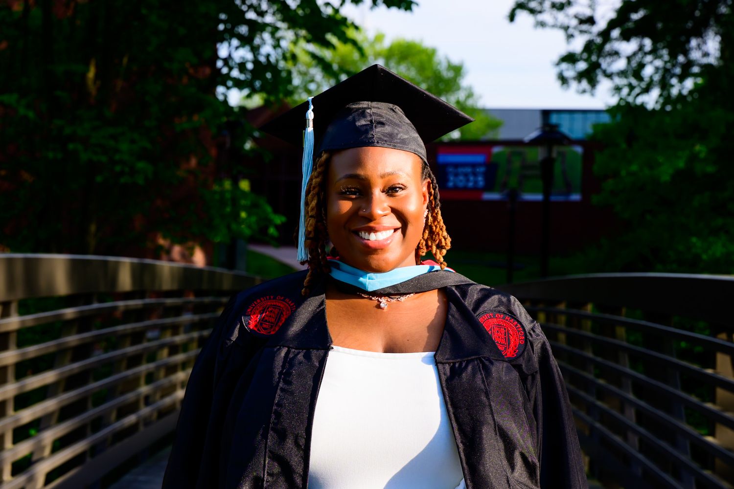 A graduate in cap and gown smiles brightly on a footbridge during a sunny graduation ceremony.