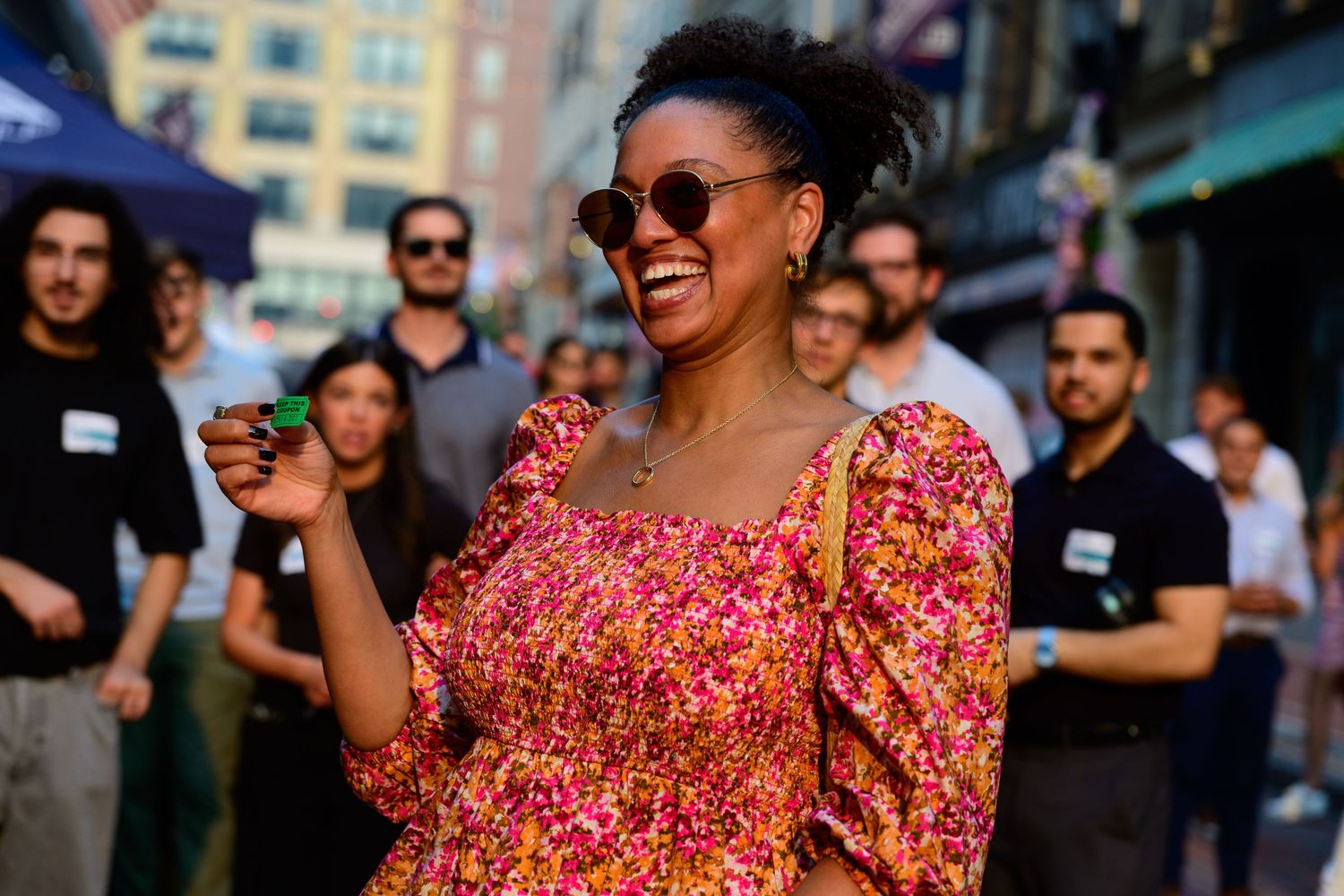 A joyful woman in a floral dress laughs outdoors on a city street surrounded by a crowd of people.