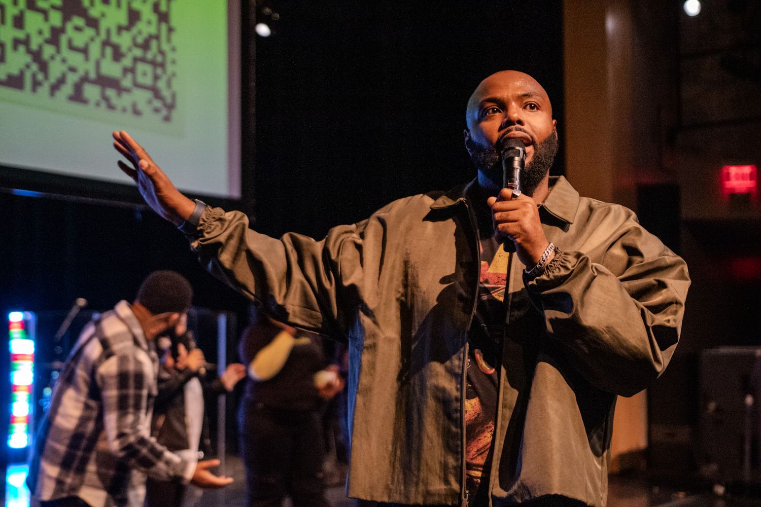 Performer in casual attire speaks into microphone on dimly lit stage with LED lighting.