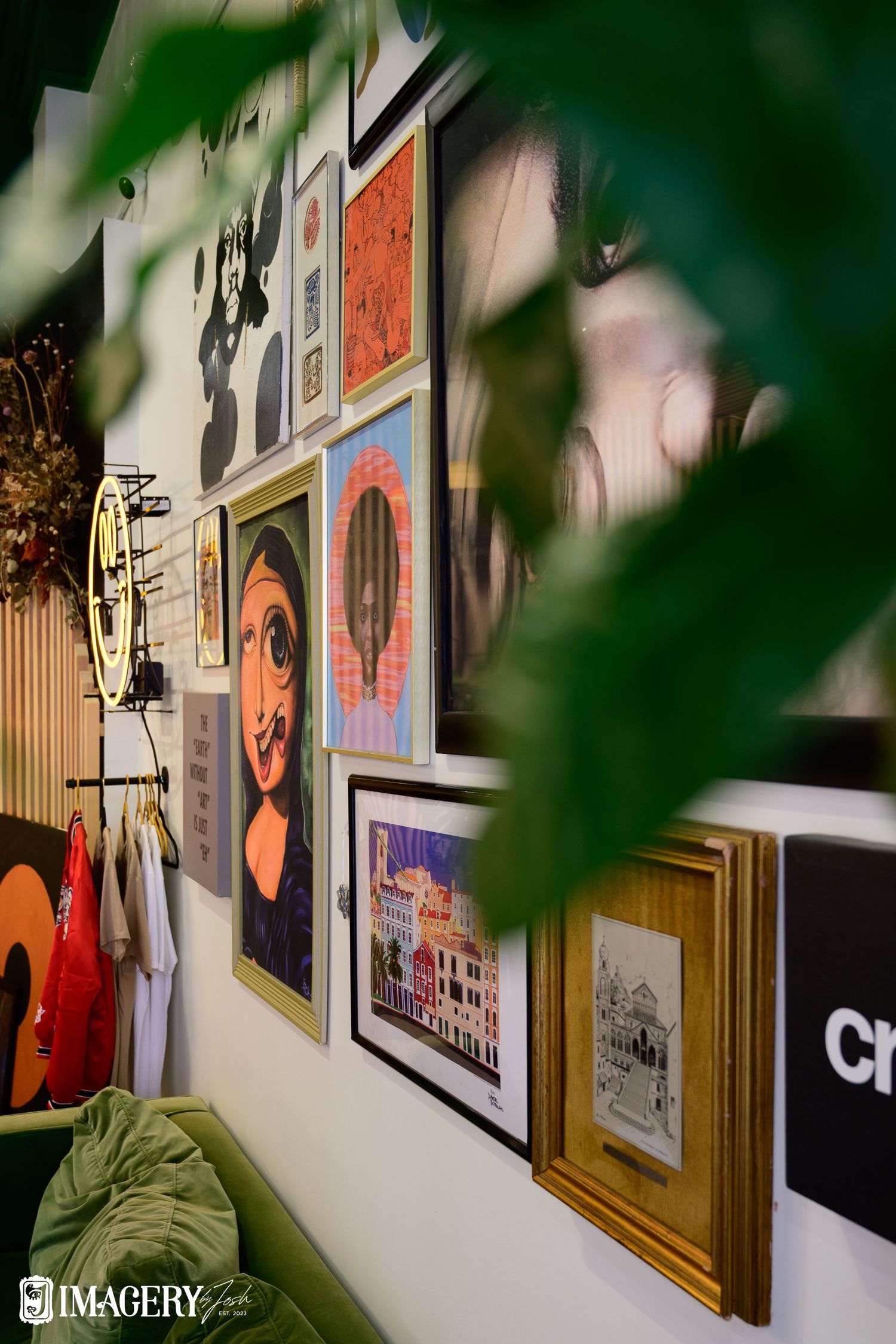 A gallery wall of framed artwork and portraits seen through green foliage with a guitar visible in the corner.
