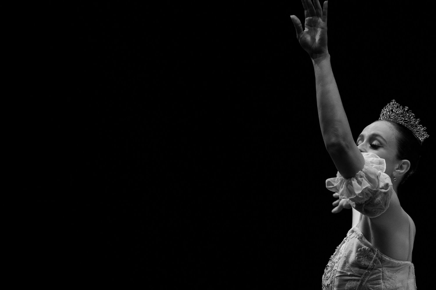 Black and white photo of a ballerina wearing a tiara, raising her arm gracefully against a dark background.