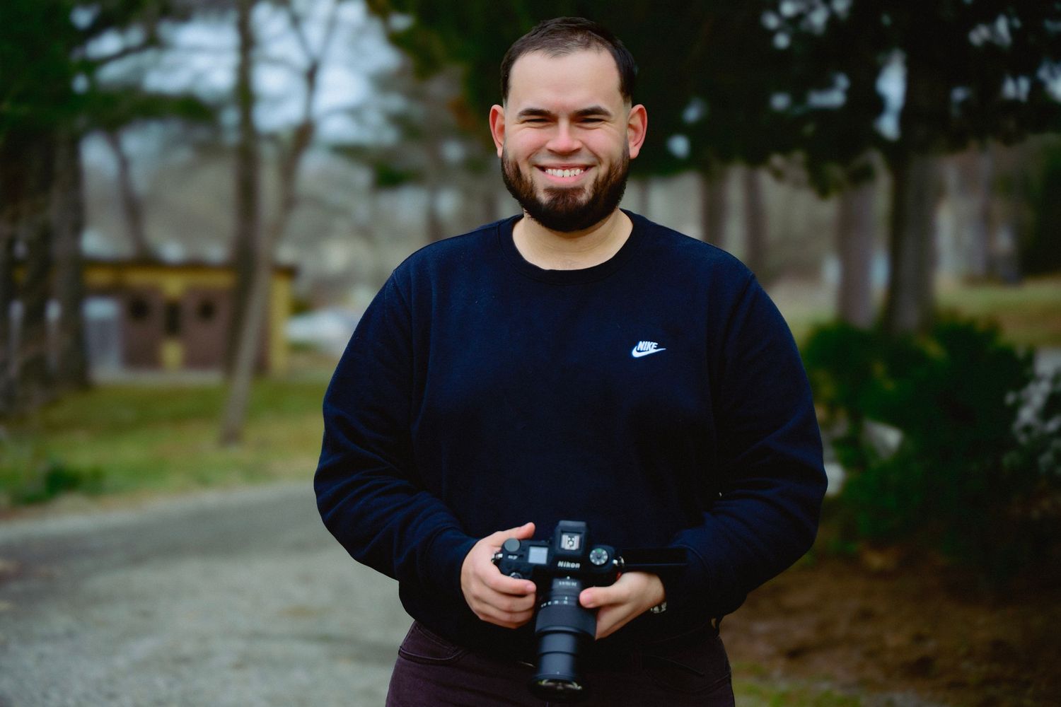 Smiling man with beard holding DSLR camera outdoors, wearing navy Nike sweatshirt, standing on path with trees behind.