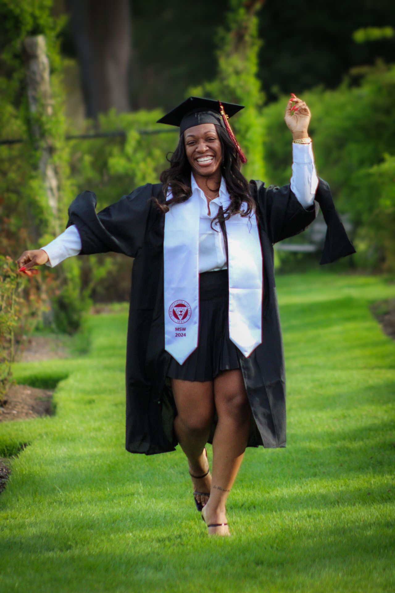 Graduate celebrating in academic regalia with arms raised joyfully on grass lawn.