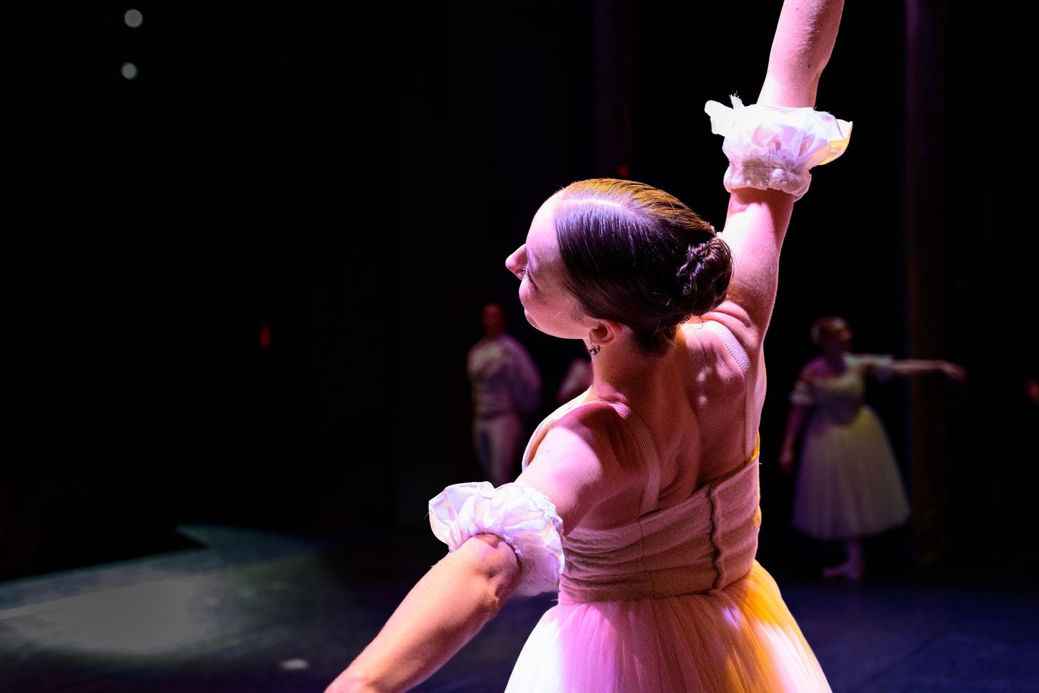A ballerina in a yellow tutu performs gracefully on stage with arm raised, with other dancers visible in the background.