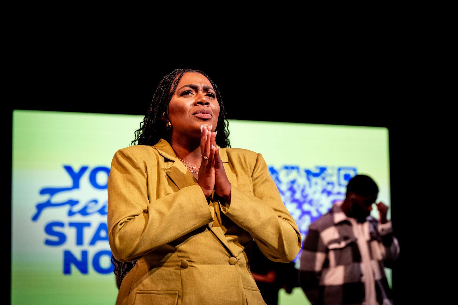 A person in a yellow blazer stands with clasped hands in prayer position against a green stage backdrop.