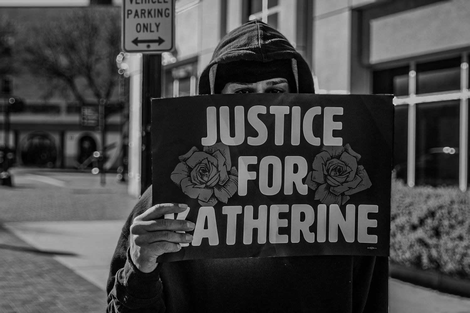 Black and white photo of protest sign reading 'Justice for Katherine' held against urban backdrop.