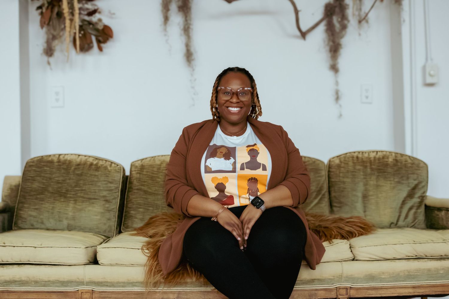 Person in brown cardigan sits on vintage couch against white wall with hanging foliage.