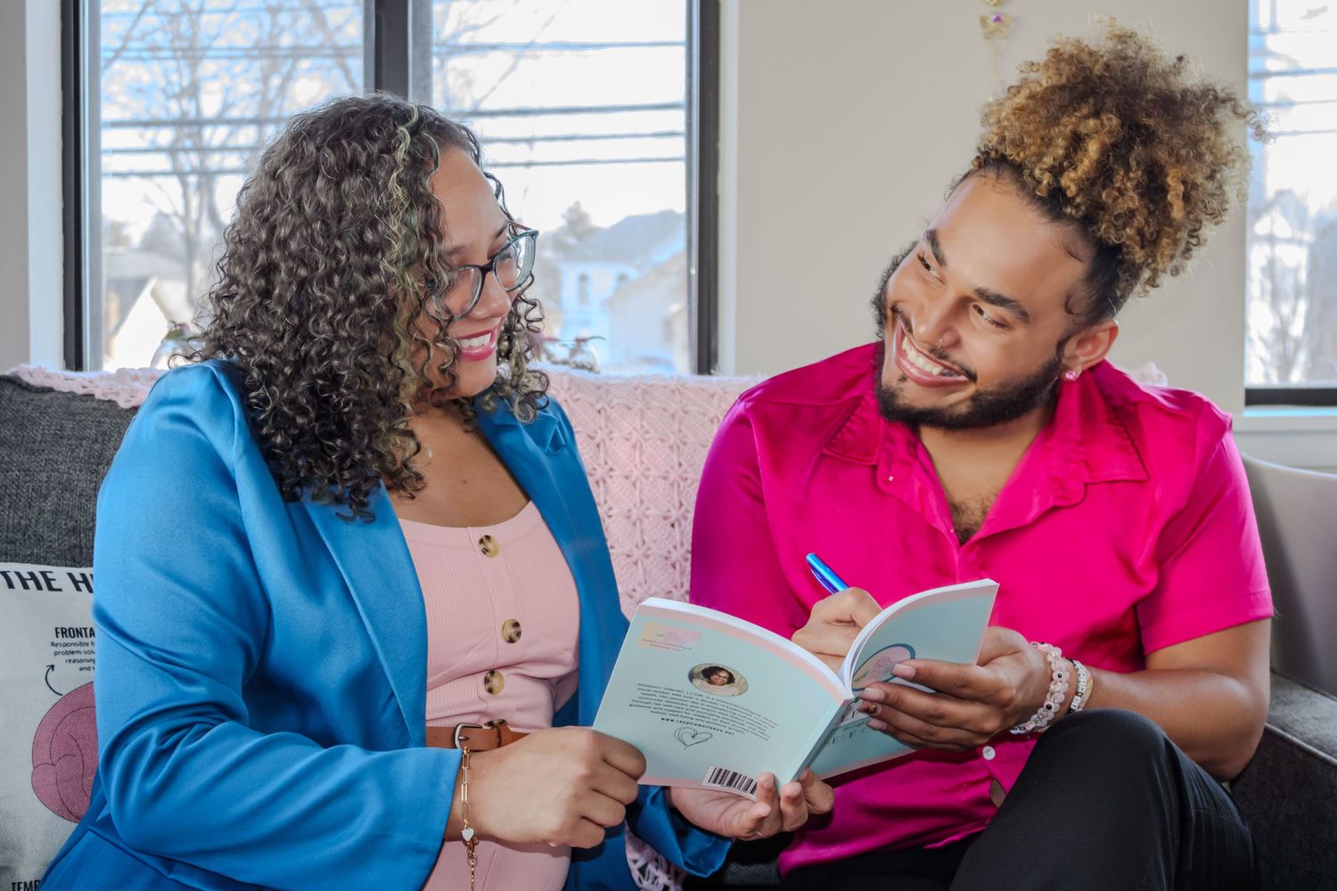 Two people sit together on a couch and smile while writing in notebooks during a casual meeting or study session.