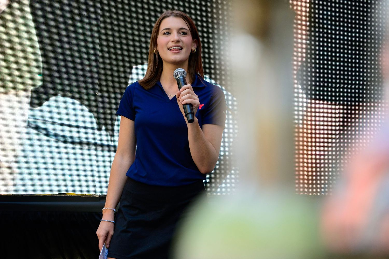 Young woman in blue top speaking into microphone on stage at outdoor event.