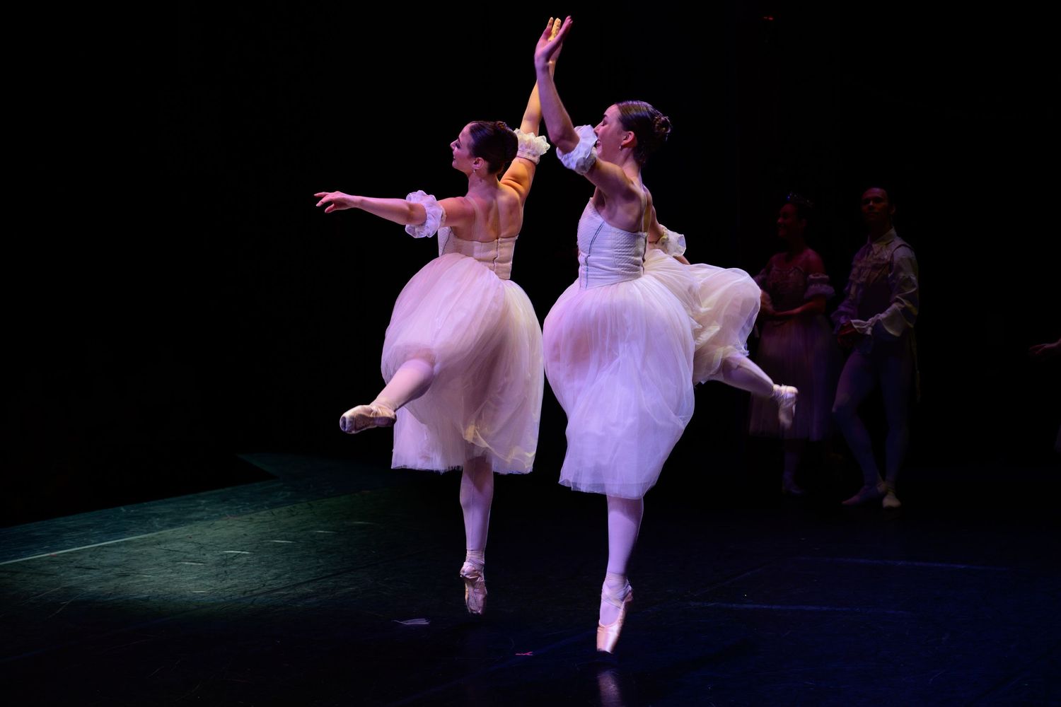 Two ballerinas in white tutus perform gracefully on stage, with a male dancer visible in the background.