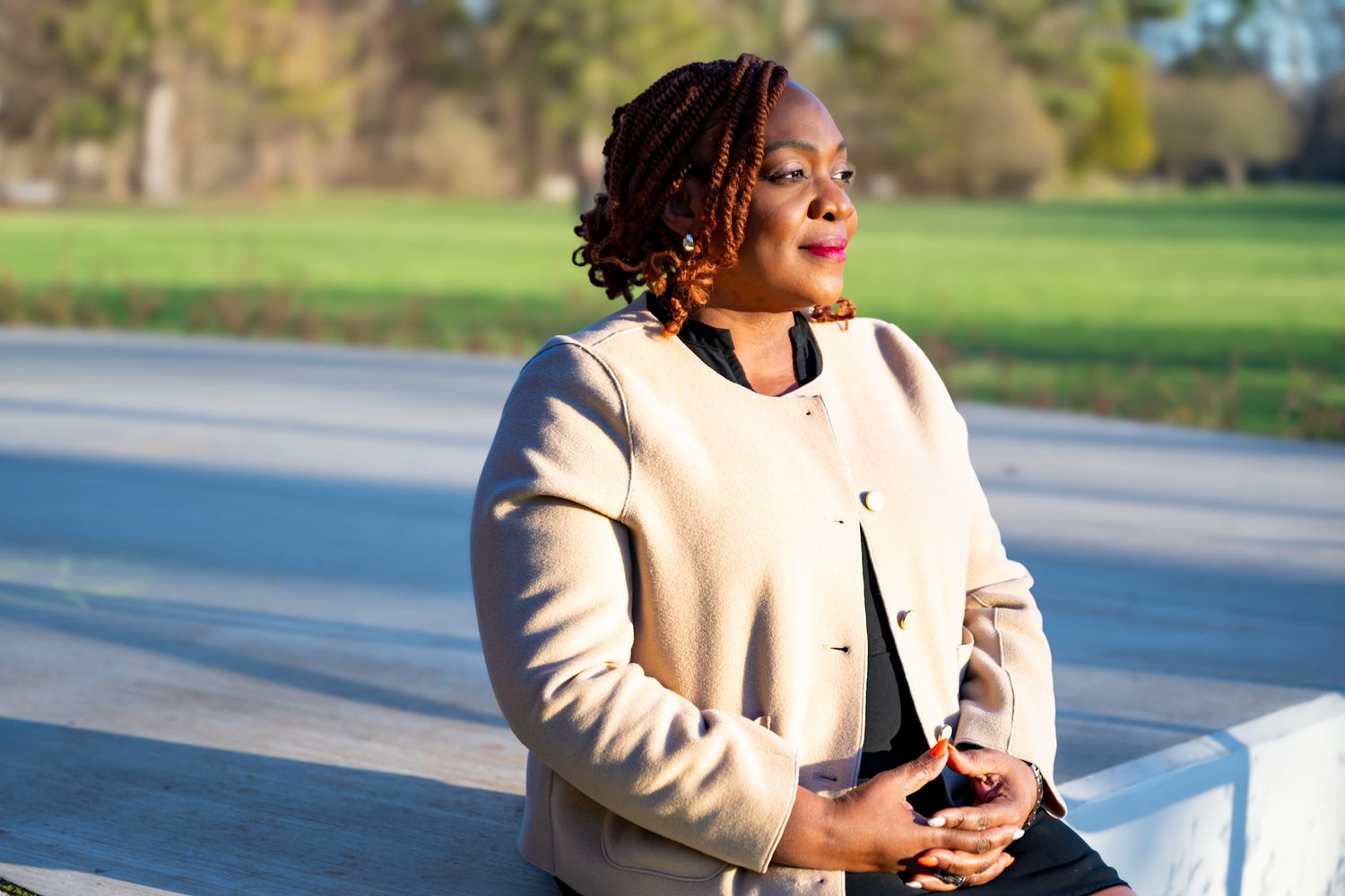 Someone wearing a cream-colored coat smiles while standing outdoors on a sunny day.