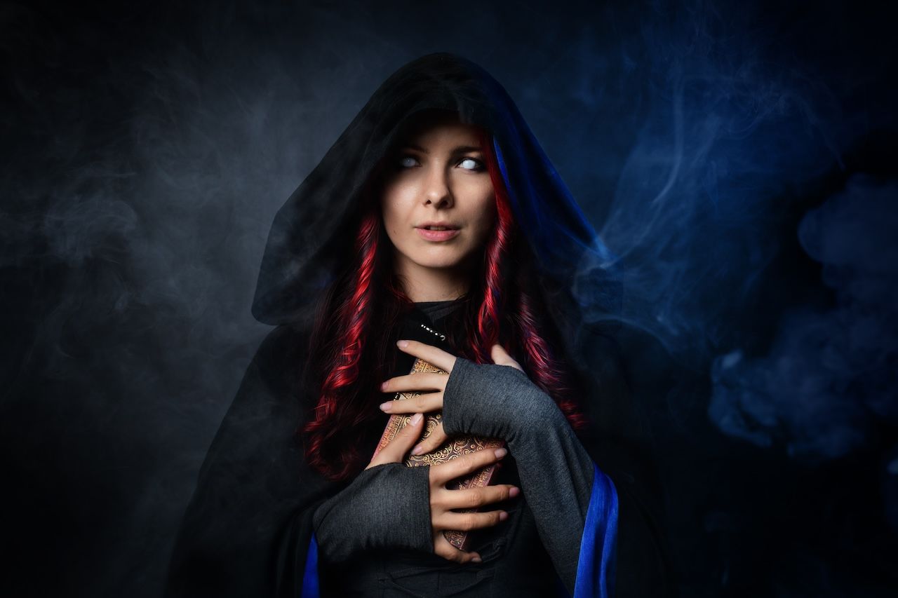Female Archetypes: Dramatic portrait of a mysterious figure in a dark hooded cloak holding a book against a smoky blue background.