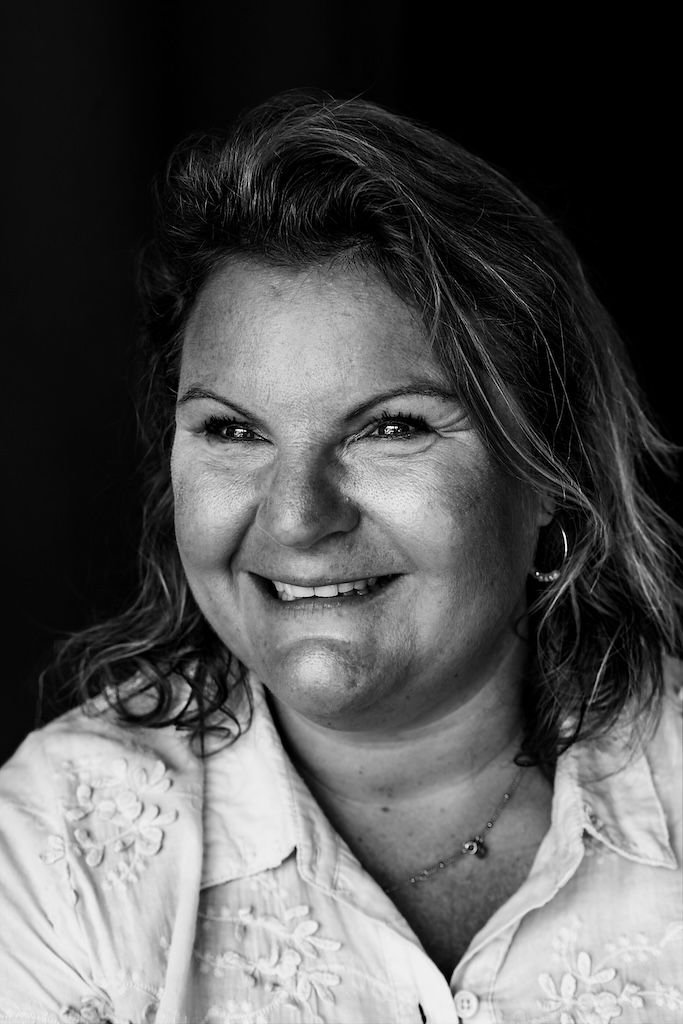 Zwart-witportret van een lachende vrouw van middelbare leeftijd met krullend haar, gekleed in een witte blouse met bloemenprint en een halsk