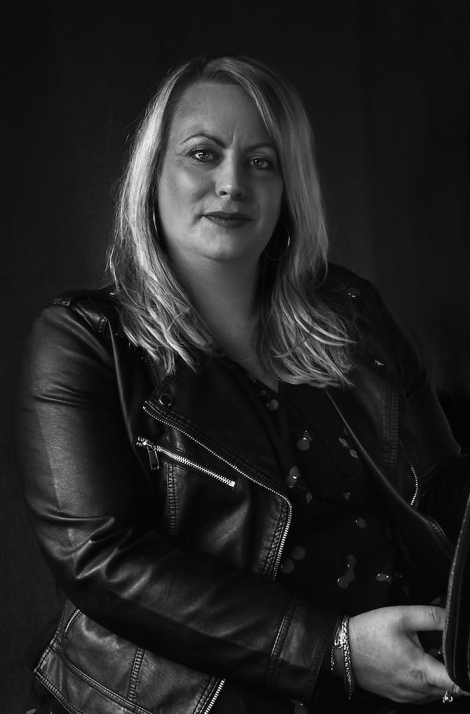 Zwart-witportret van een blonde vrouw in een leren jasje, die zelfverzekerd poseert tegen een donkere achtergrond.