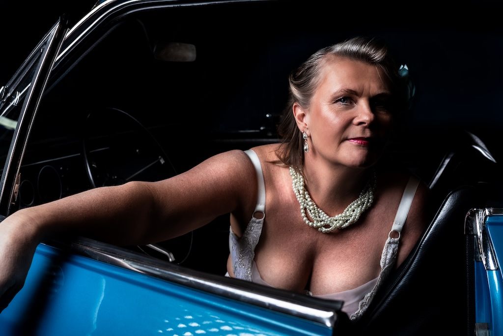 Woman in low-cut top and pearl necklace leaning against a classic blue car in dramatic lighting.