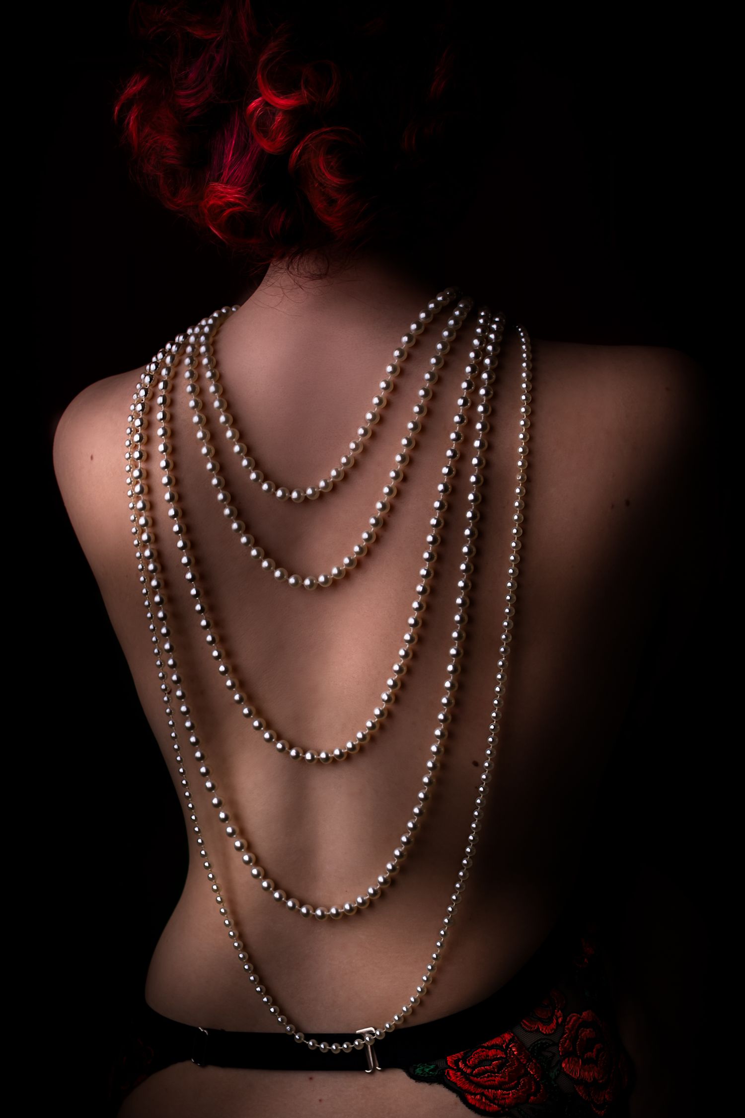 Artistieke boudoirfoto van een naakte rug versierd met gedrapeerde parelkettingen tegen een donkere achtergrond met rode accenten.
