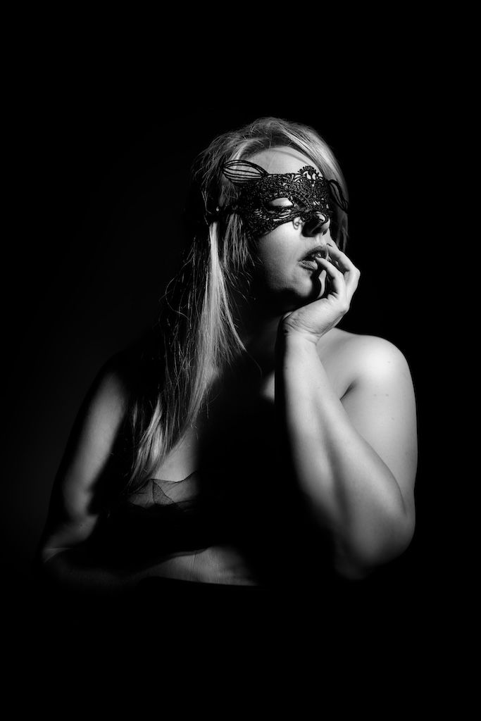 Boudoirfoto van een vrouw met een kanten masker tegen een donkere achtergrond in sfeervol licht.