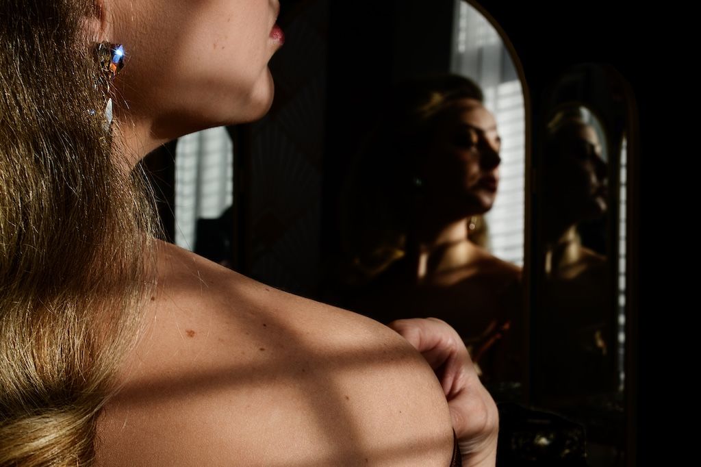 Woman with earring and bare shoulder stands before mirror reflecting a man in dramatic light.