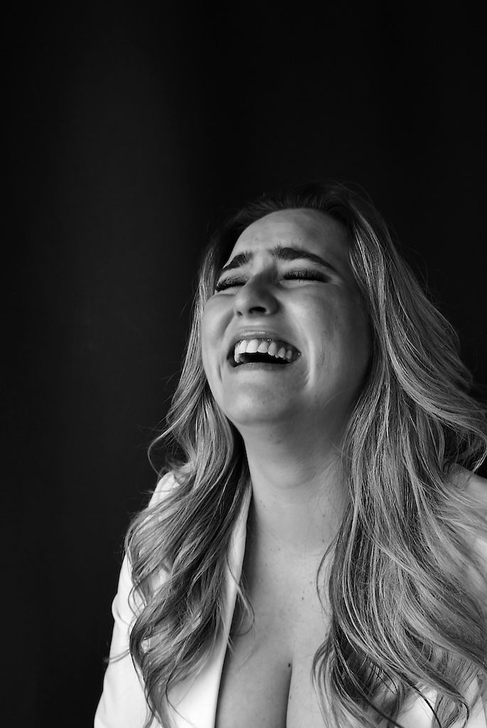 Zwart-witportret van een lachend persoon met gesloten ogen en golvend blond haar tegen een donkere achtergrond.