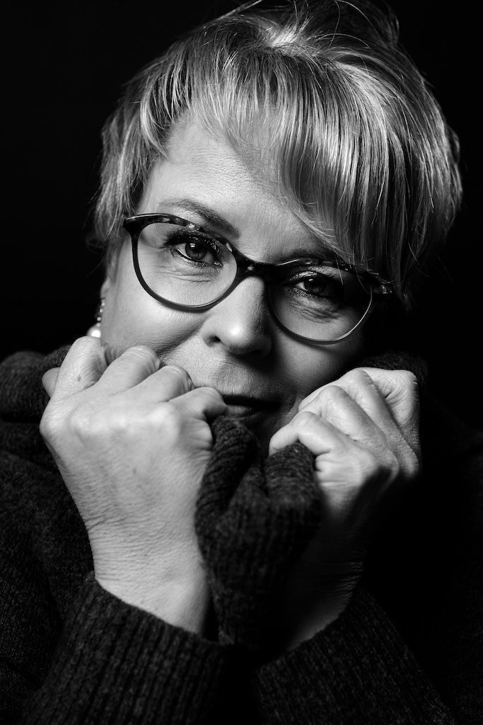 Zwart-witportret van een vrouw met een bril die haar coltrui tot aan haar gezicht optrekt, tegen een donkere achtergrond.