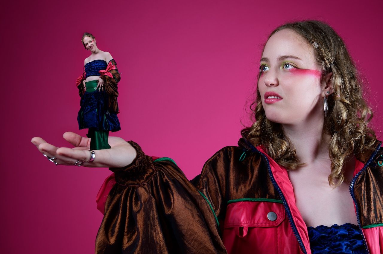 A woman in Victorian-style dress holds a miniature version of herself against a pink background.