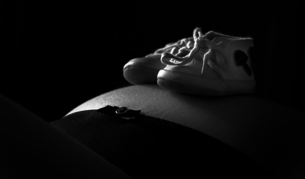 Zwart-wit zwangerschapsfoto van babyschoentjes op een buik tegen een donkere achtergrond. Zeewolde