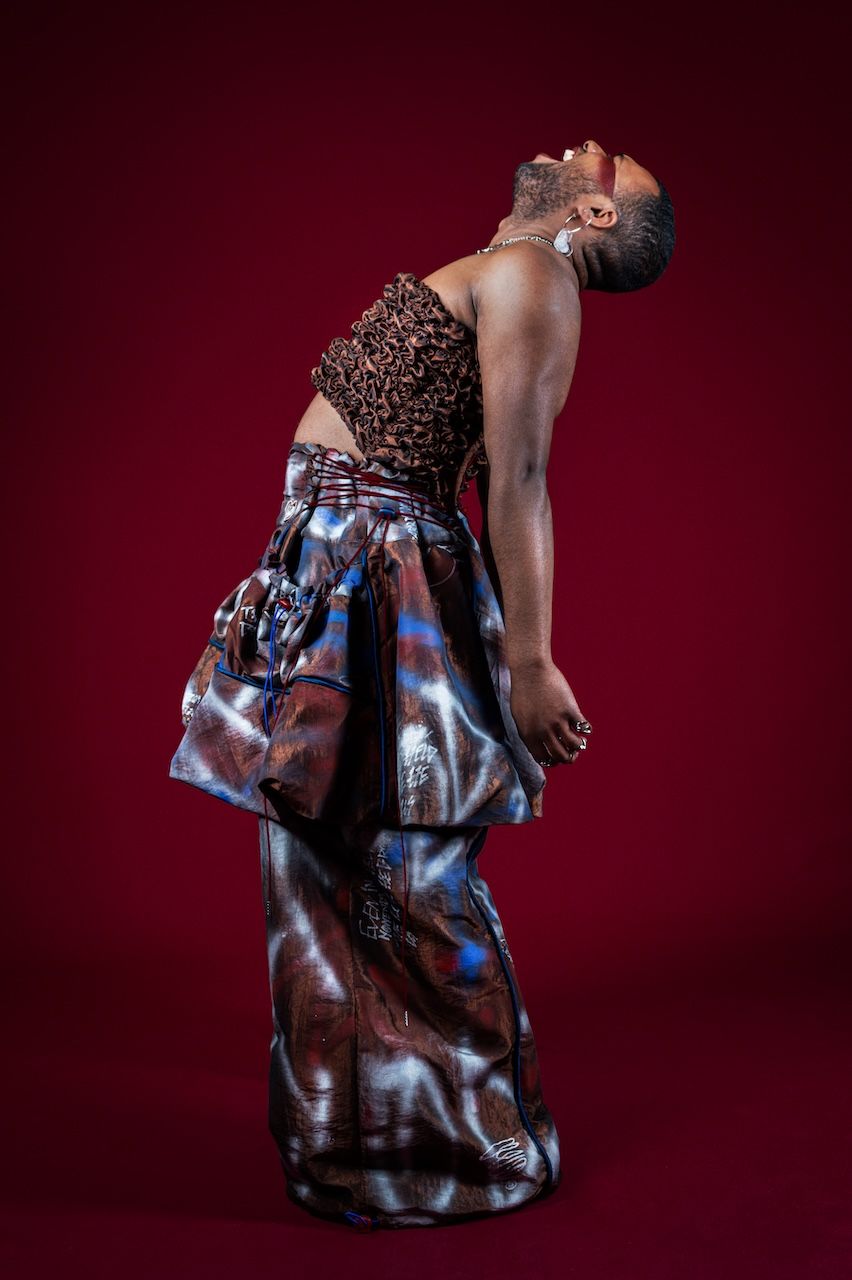 A figure in a leopard print top and flowing blue patterned skirt poses dramatically against a deep red background.