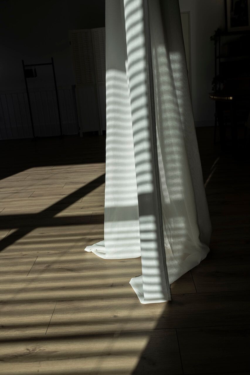 Zonlicht dat door de jaloezieën naar binnen valt, werpt gestreepte schaduwen op de witte gordijnen en de houten vloer in fotostudio Zeewolde