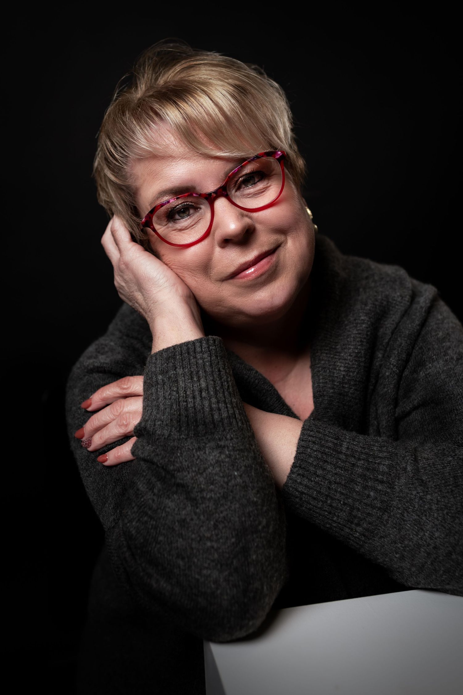 Studioportret tegen zwarte achtergrond van een vrouw met rode bril en donkere trui in warm licht.