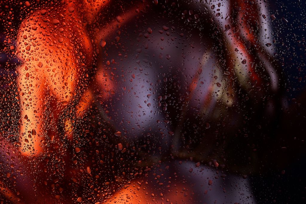 Close-up van regendruppels op glas met levendige rode en oranje  licht op het bovenlichaam van een vrouw.