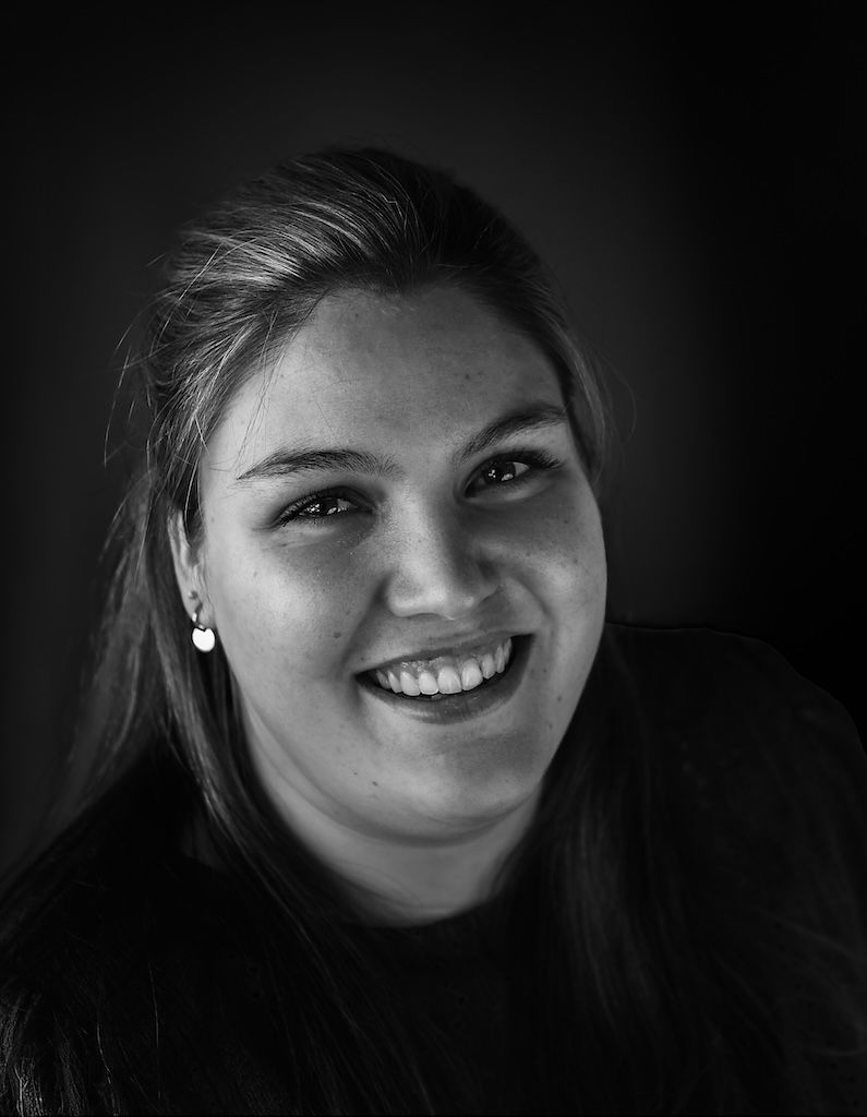 Zwart-witportret van een lachende jonge vrouw met lang haar en kleine oorbellen, tegen een donkere achtergrond.