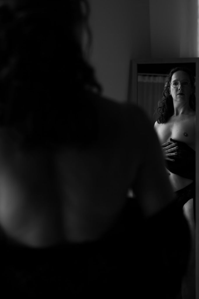 Zwart-witfoto van een vrouw die zichzelf in een spiegel bekijkt, gedeeltelijk ontkleed in een schemerig verlichte ruimte. Boudoirfotoshoot