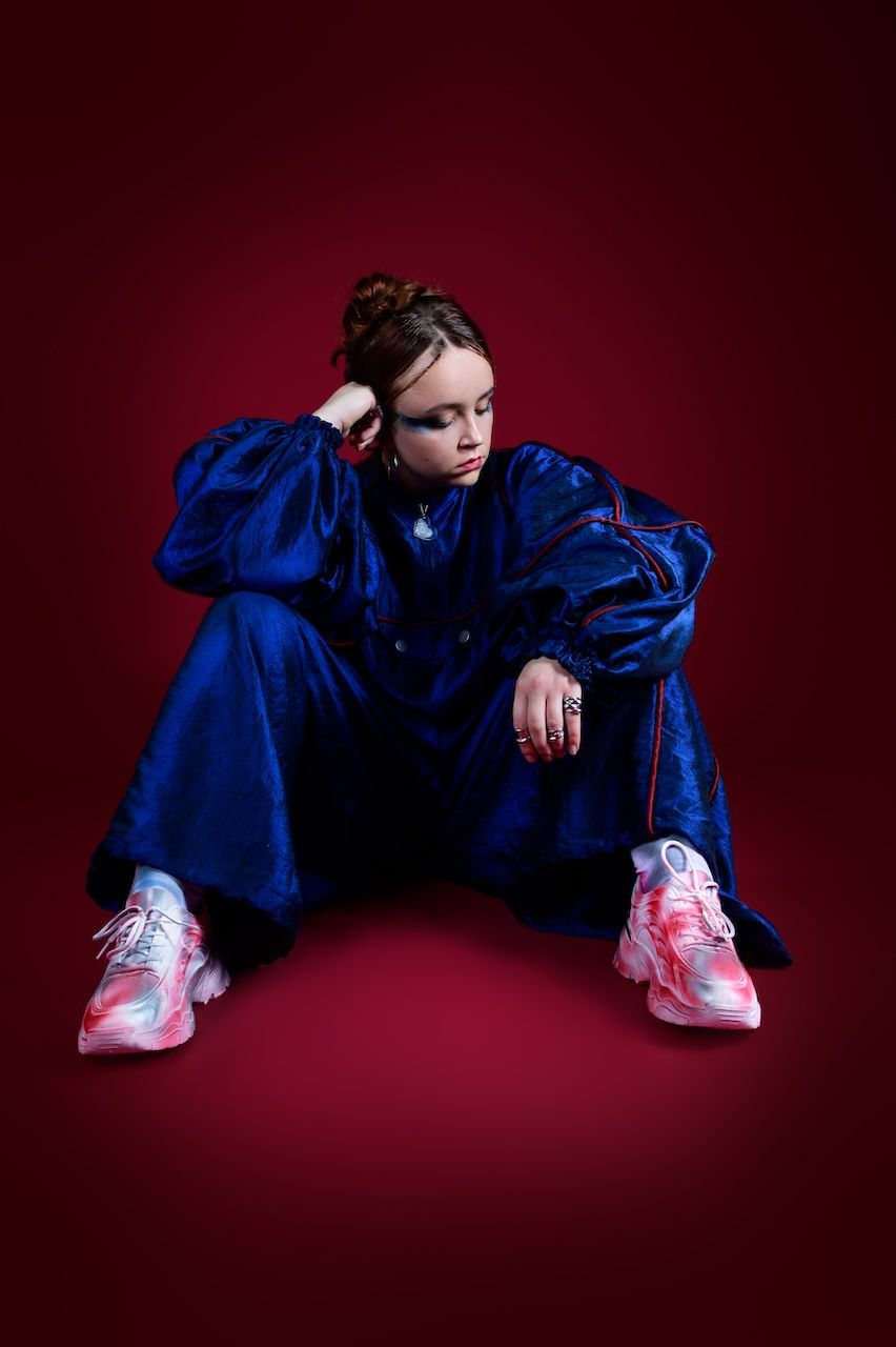 Person in bright blue athletic wear and pink sneakers poses dramatically against dark red studio backdrop.
