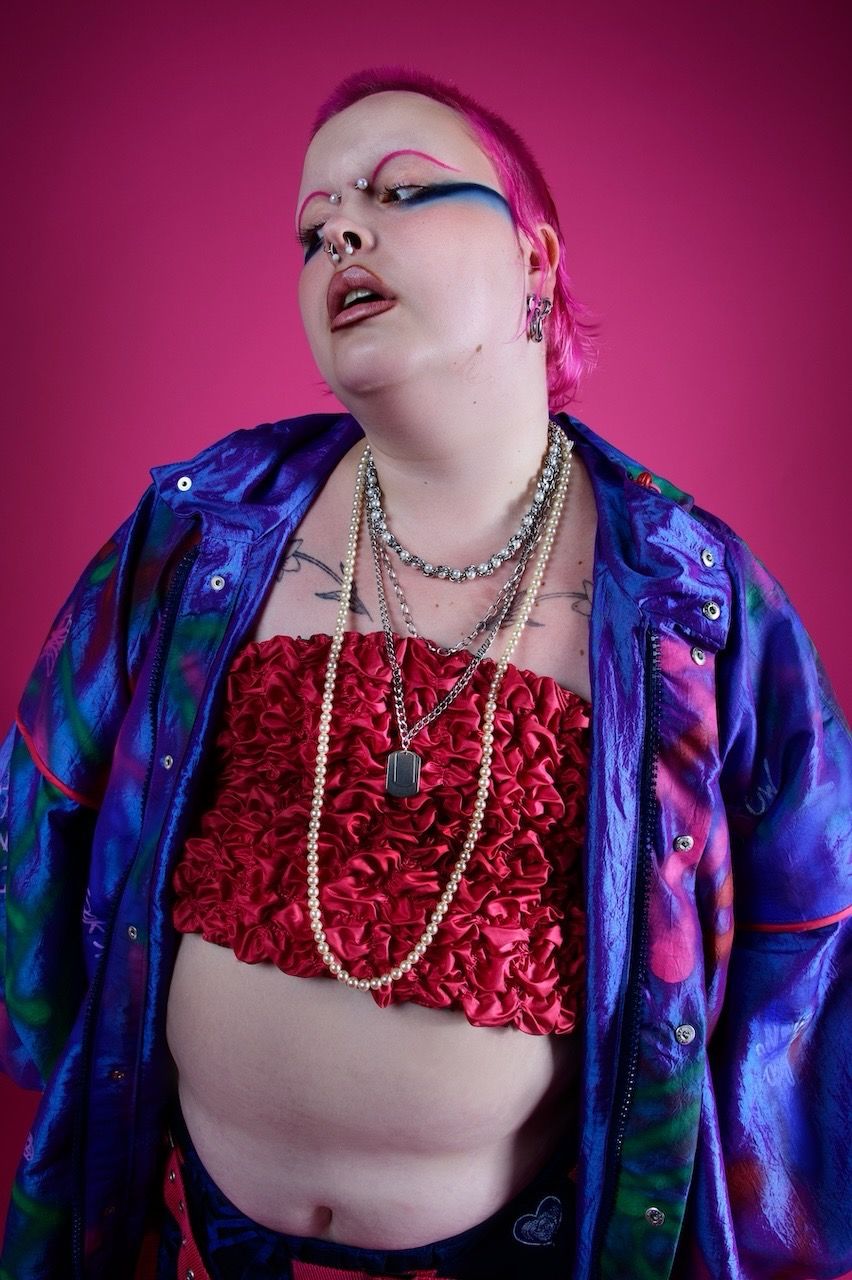 Fashion portrait with red crop top, blue iridescent jacket and pearl necklaces against pink background.