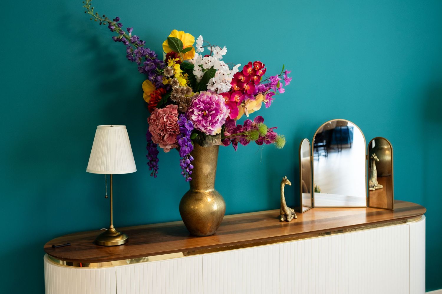 Kleurrijk bloemstuk in een messing vaas tegen een turquoise muur, met een houten dressoir en een witte tafellamp, fotostudio Zeewolde